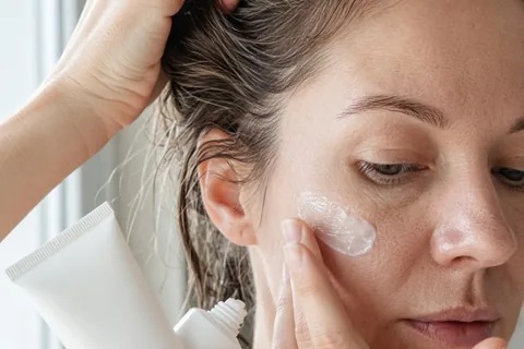 Close-up of a hand applying azelaic acid cream to a woman's cheek with the product tube visible