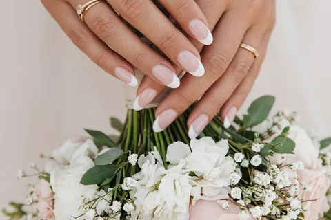 Close-up of a bride's hands with a soft baby boomer French manicure holding a bouquet of flowers, with wedding rings on the ring fingers.