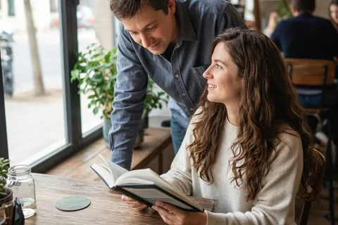 A man in his thirties with short brown hair wearing a light blue button-up shirt talking to a woman in her mid-twenties with long wavy brown hair wearing a simple beige sweater at a wooden table in a 