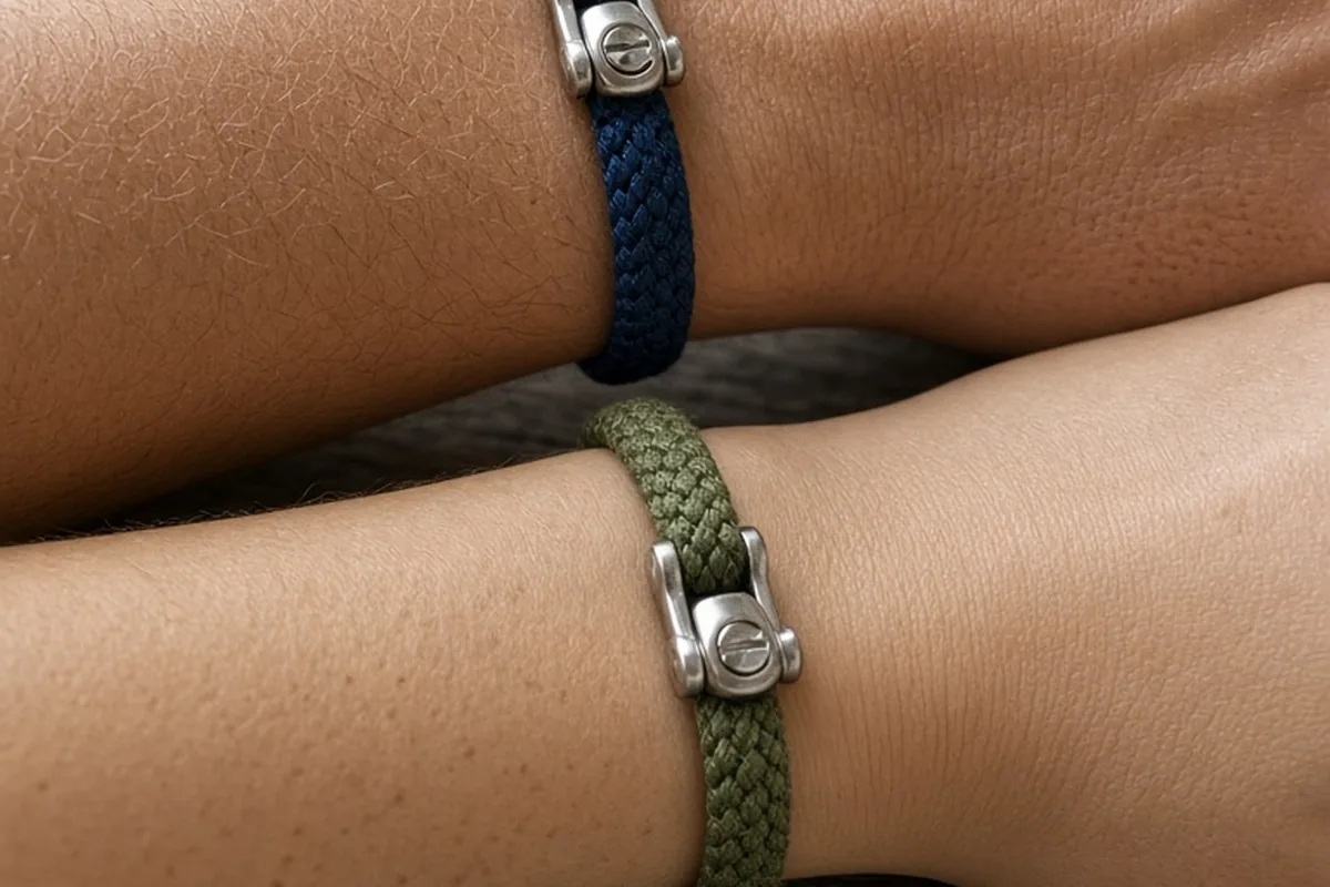 Two adult wrists side by side wearing flat braided marine cord bracelets with stainless steel screw-pin clasps on a wooden table under soft natural light.